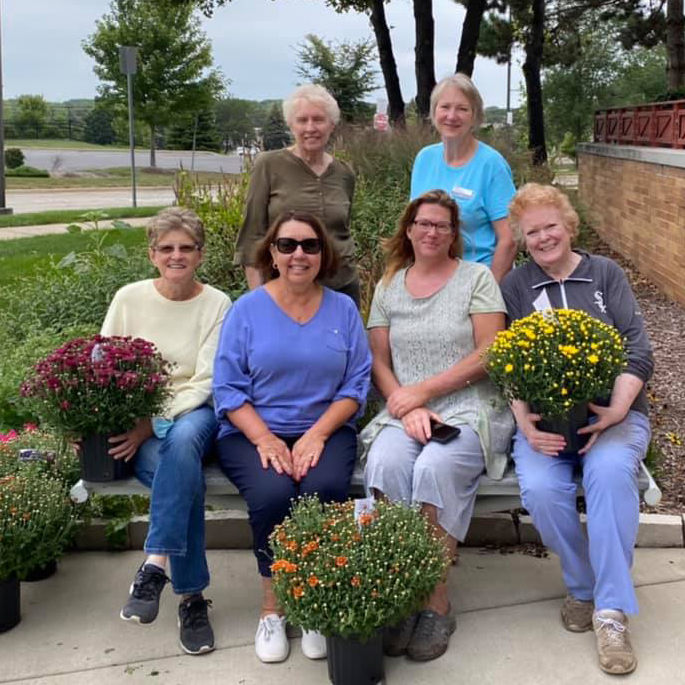 6 Friends of the Library members sitting on an outdoor bench with pots of mums