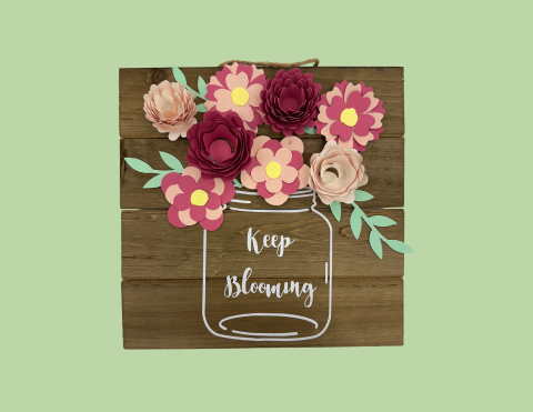 An image of vinyl in the shape of a mason jar with the text "Keep Blooming". There are multi-colored paper flowers glued at the top of the wooden sign.