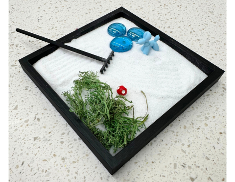 The image shows a box filled with sand, a miniature rake, pebbles, fake moss, and a balloon dog figure.